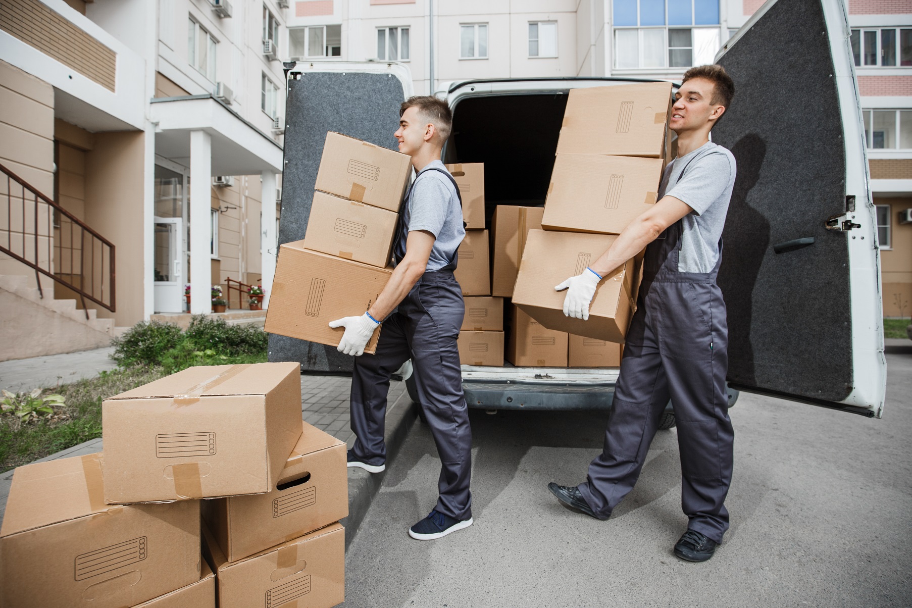 Two young handsome smiling workers wearing uniforms are unloading the van full of boxes. There is a block of flats in the background.